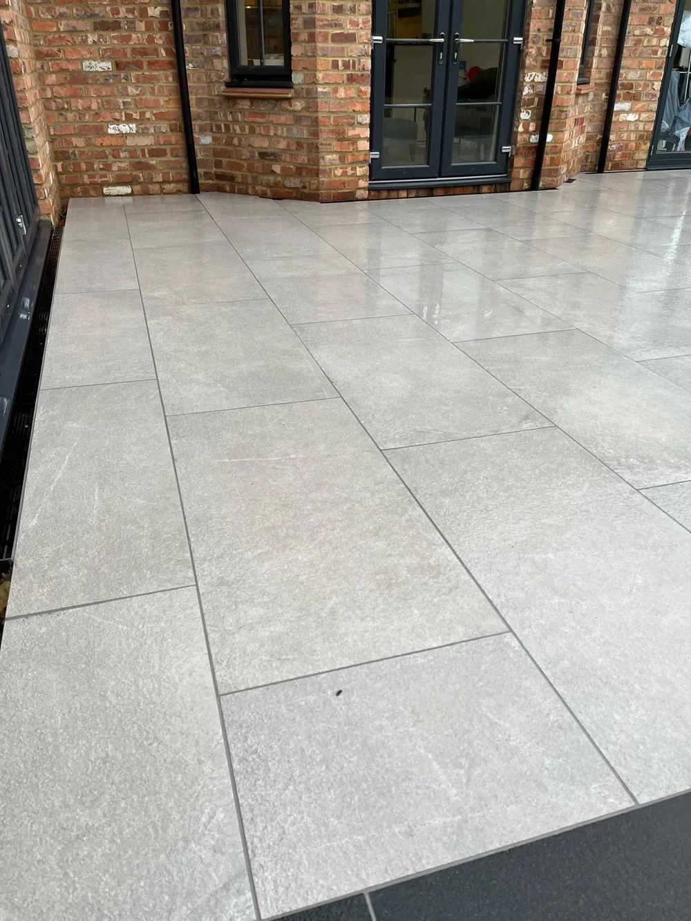 Grey porcelain patio laid tight to dark framed doors at the back of a brick house