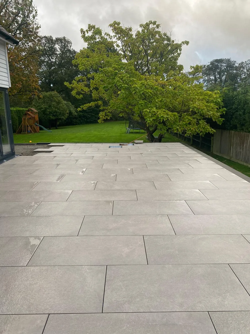 Wide grey porcelain patio running from the house to a lawn under mature trees