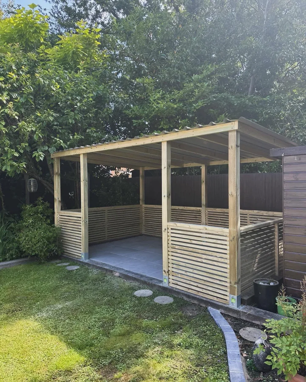 Timber garden shelter with slatted sides and a paved floor on a lawn