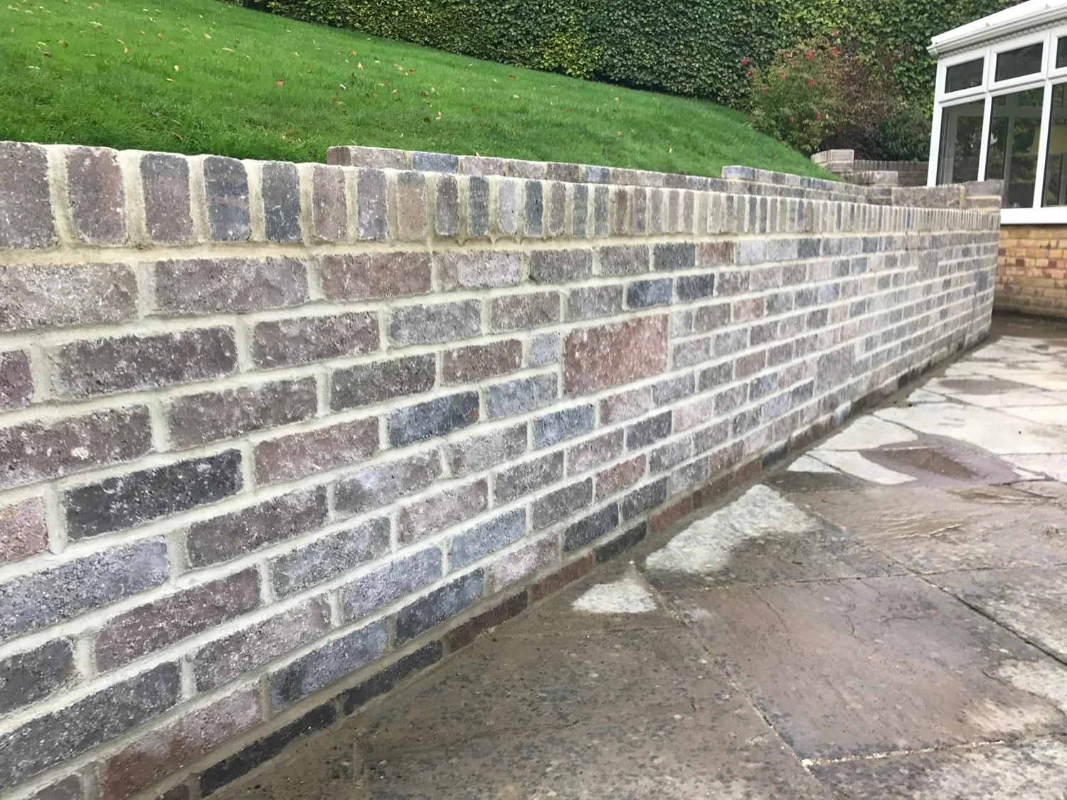 Reclaimed brick retaining wall holding a lawn above a sandstone patio beside a conservatory